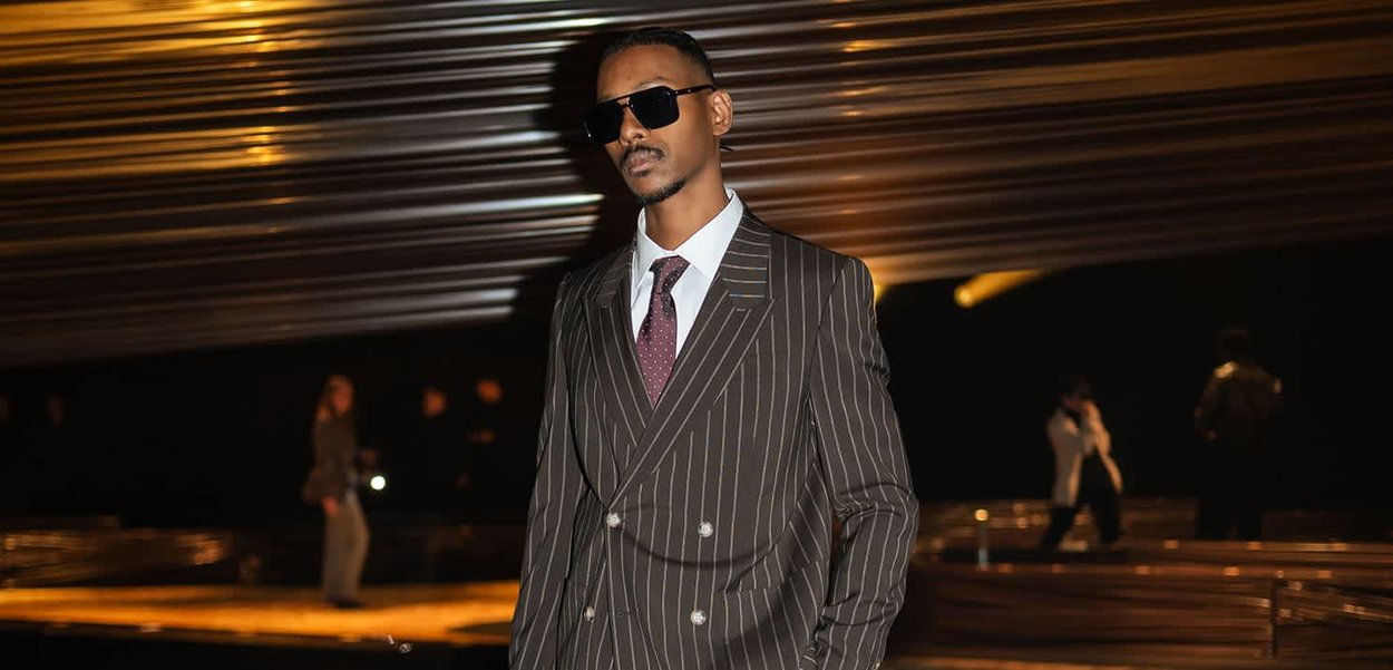 Hombre con traje oscuro de rayas, camisa blanca, corbata burdeos y gafas de sol, de pie bajo un techo moderno revestido de madera, con personas desenfocadas al fondo.