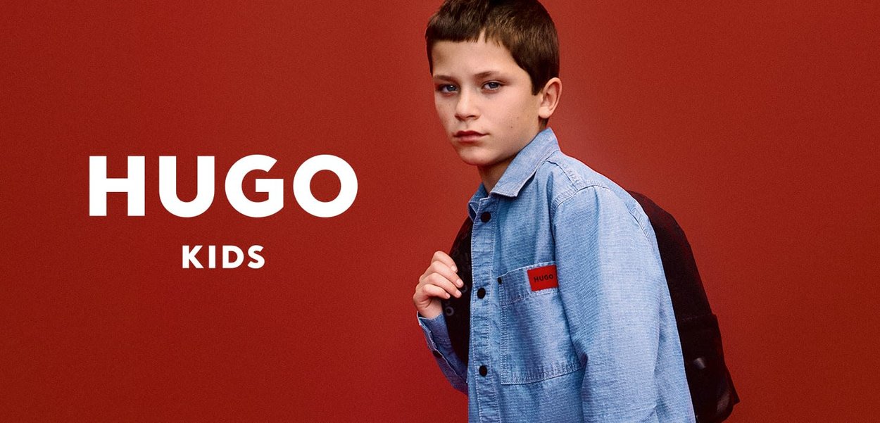 Light blue denim jacket with a red label, black backpack, against a solid red background. Boy with short brown hair looks directly at the camera.