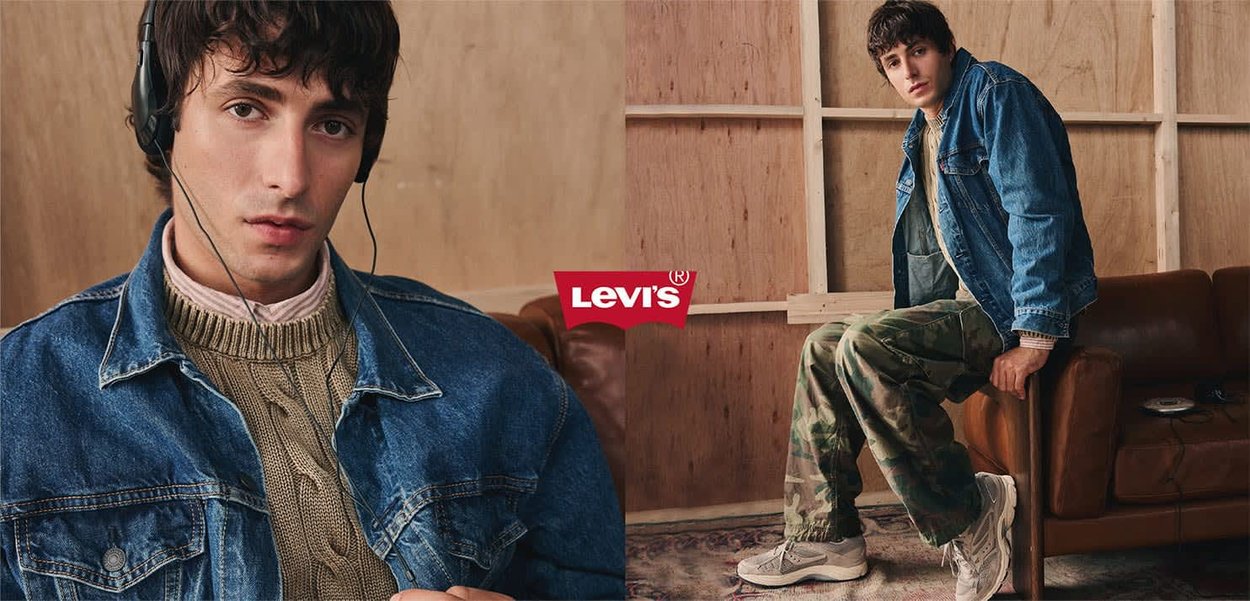 Junger Mann trägt Jeansjacke, Strickpullover und Tarnhosen, sitzt auf einem Stuhl in einem hölzernen Raum mit braunem Ledersofa.