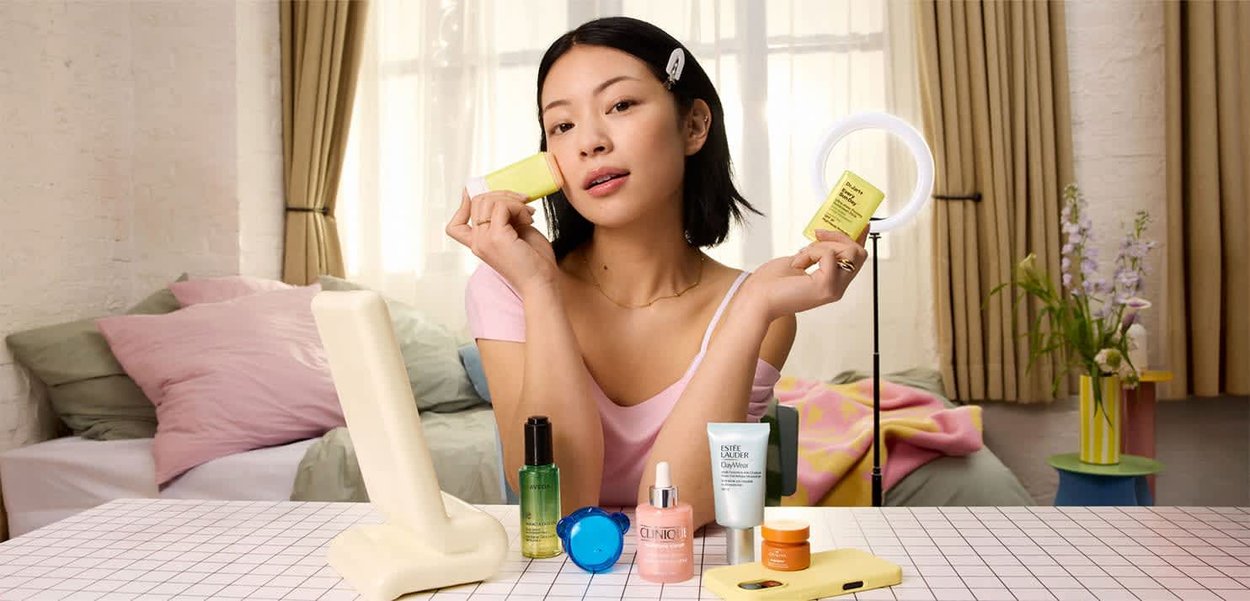 Junge Frau, die gelbe Hautpflegeprodukte an einem Tisch mit verschiedenen Schönheitsartikeln und einem Ringlicht in einem hellen Schlafzimmer vorführt.