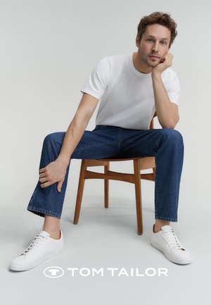 Weisses Rundhals-T-Shirt, blaue Jeans und weisse Sneaker mit braunen Akzenten, sitzend auf einem Holzstuhl vor einem schlichten Hintergrund.