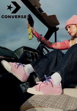 Personne portant des baskets Converse roses, des chaussettes blanches et un bonnet rose, assise dans une voiture, réglant le rétroviseur avec un désodorisant suspendu.