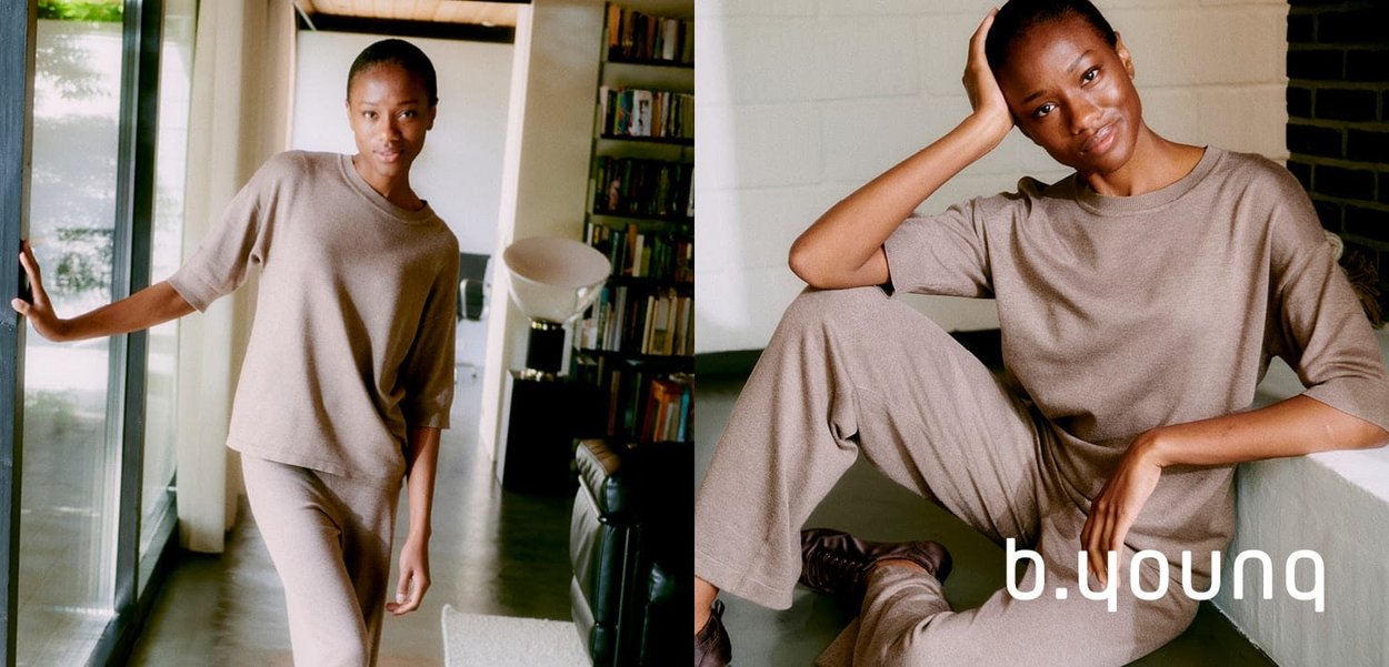 Una modella indossa un completo in maglia beige a tinta unita, caratterizzato da una maglietta a maniche corte e pantaloni larghi, sullo sfondo di un ambiente interno.