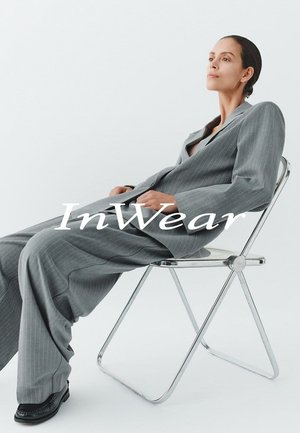 Grau gestreifter Anzug mit weitem Schnitt und leicht übergroßer Passform, getragen im Sitzen auf einem chromfarbenen Klappstuhl vor weißem Hintergrund.