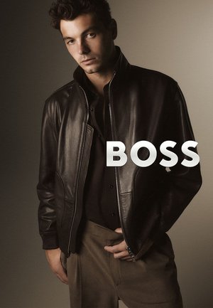 Giacca di pelle marrone con collo alto, chiusura con cerniera e tasche con cerniera. Modello con maglia scura e pantaloni chiari, con sfondo neutro.