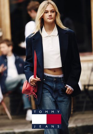 Femme blonde marchant en plein air portant un blazer bleu marine, un polo blanc court, un jean bleu, et tenant un sac à main rouge, avec des personnes floues en arrière-plan.