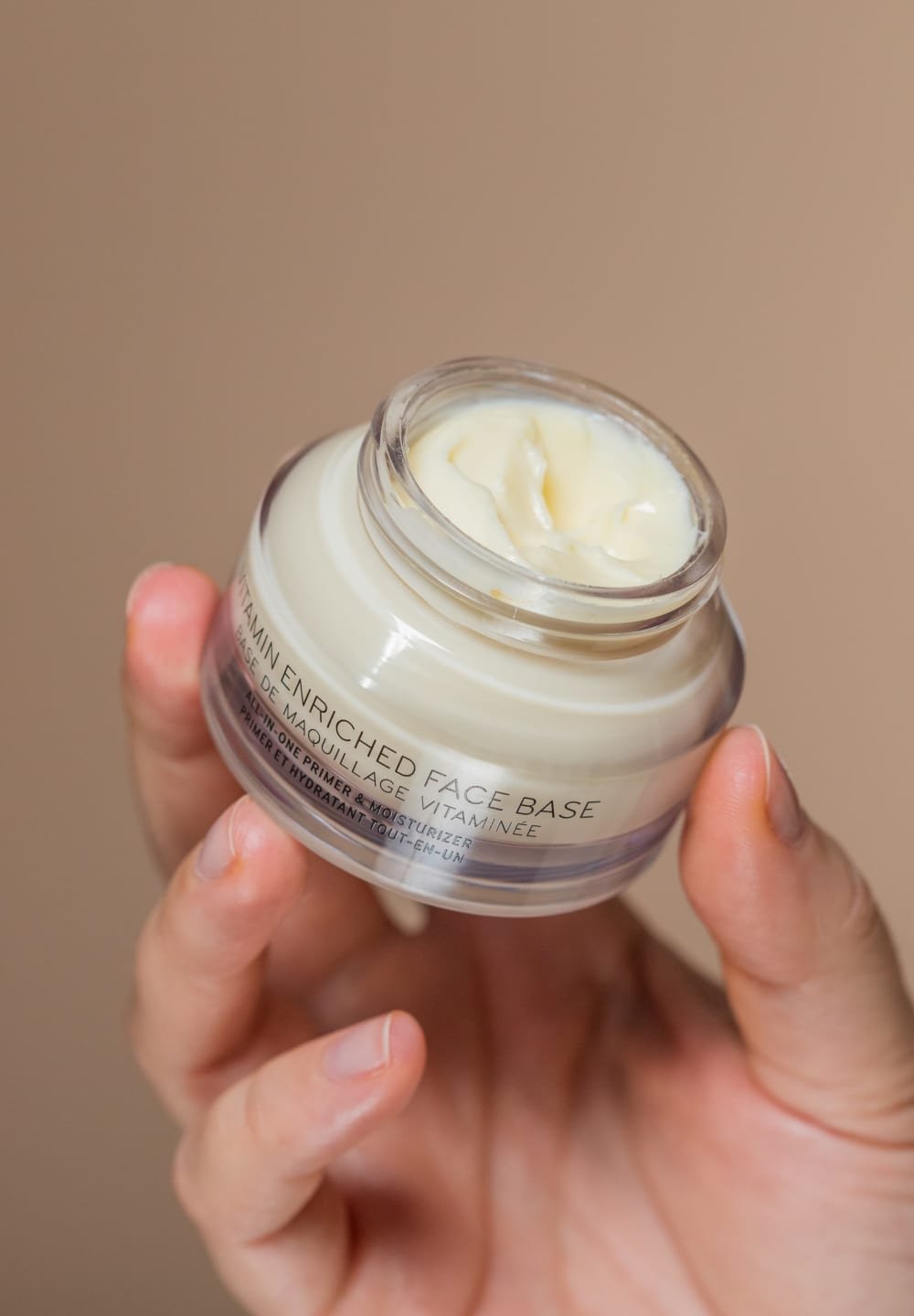 Klart glasburk med en cremet, beige ansigtsmoisturizer. Etiketten siger "Vitamin Beriget Ansigtsbase" med sort tekst. Glat tekstur synlig.