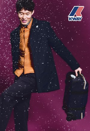 Abrigo negro sobre una camisa marrón, sosteniendo una maleta negra con ruedas. Fondo con copos de nieve cayendo sobre un fondo púrpura. Presencia del logo de K-Way.