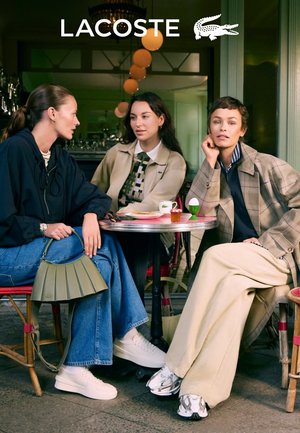Trois femmes sont assises à une petite table ronde. Elles portent des tenues tendance, dont un sweat-shirt oversize bleu, une veste en tartan beige et un pantalon large.