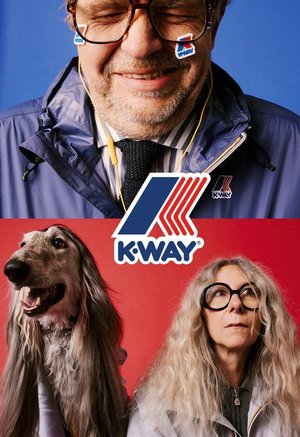 Lächelnder Mann in blauer Jacke mit K-Way-Aufklebern im Gesicht und Frau mit großen Brillen und langen Haaren neben einem langhaarigen Hund, K-Way-Logo zentriert.