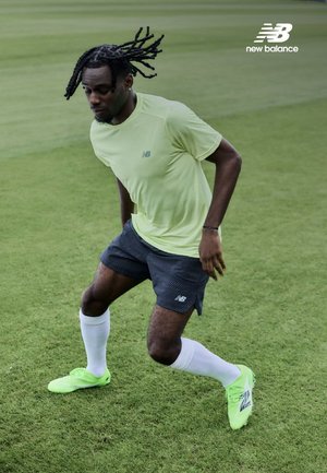 Camiseta deportiva verde lima, pantalones cortos azul marino con estampado, calcetines blancos y zapatillas verde brillante. Sujeto en movimiento en un campo de hierba. Marca New Balance visible.