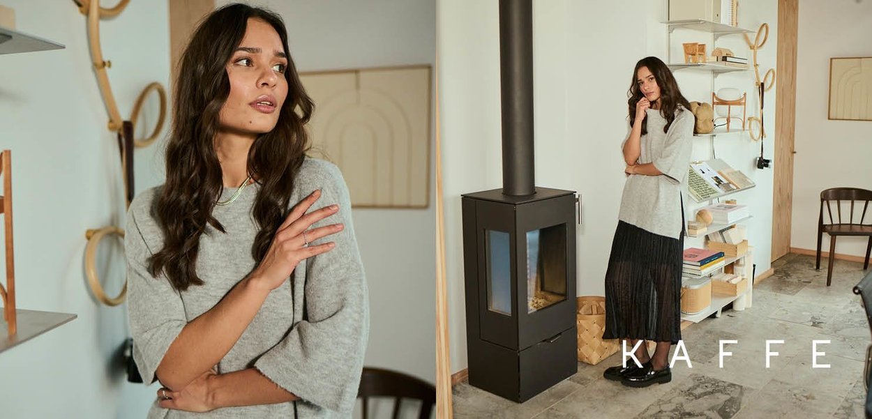 Gray oversized sweater with short sleeves, paired with a black pleated skirt. She stands near a black stove in a minimalist room.