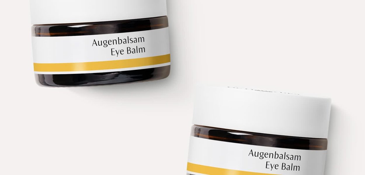 Brown glass jars with white lids; labels read "Augenbalsam Eye Balm" in black, accented by a yellow stripe at the bottom.