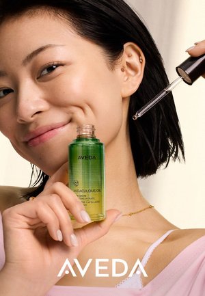 Femme aux cheveux courts et foncés tenant un flacon vert d'huile Aveda Miraculous, utilisant une pipette pour appliquer le produit près de sa joue, souriant doucement.