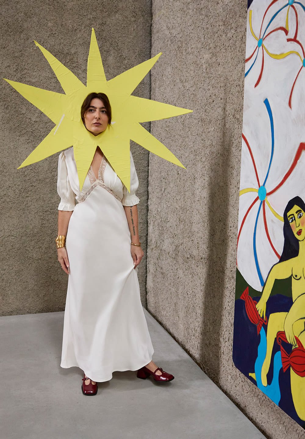Άτομο σε λευκό φόρεμα και κόκκινα παπούτσια που φορά ένα μεγάλο κίτρινο καπέλο σε σχήμα ήλιου, στέκεται κοντά σε έναν πολύχρωμο αφηρημένο πίνακα.