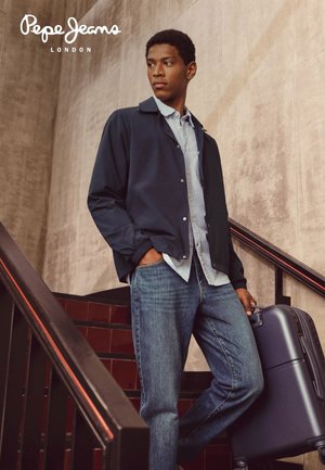 Ragazzo giovane con camicia casual, giacca blu navy e jeans in piedi sulle scale con una valigia, davanti a un muro di cemento con il logo Pepe Jeans London.