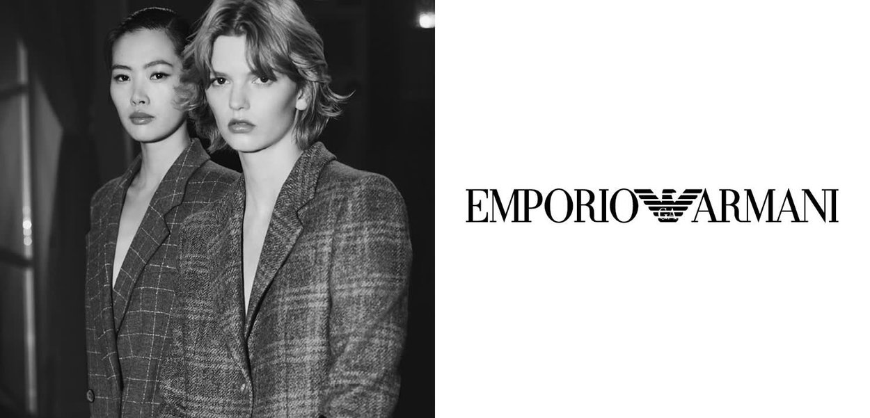 Dos modelos llevan blazers ajustados de cuadros grises. El fondo es oscuro, contrastando con la ropa clara. Logo de "EMPORIO ARMANI" a la derecha.