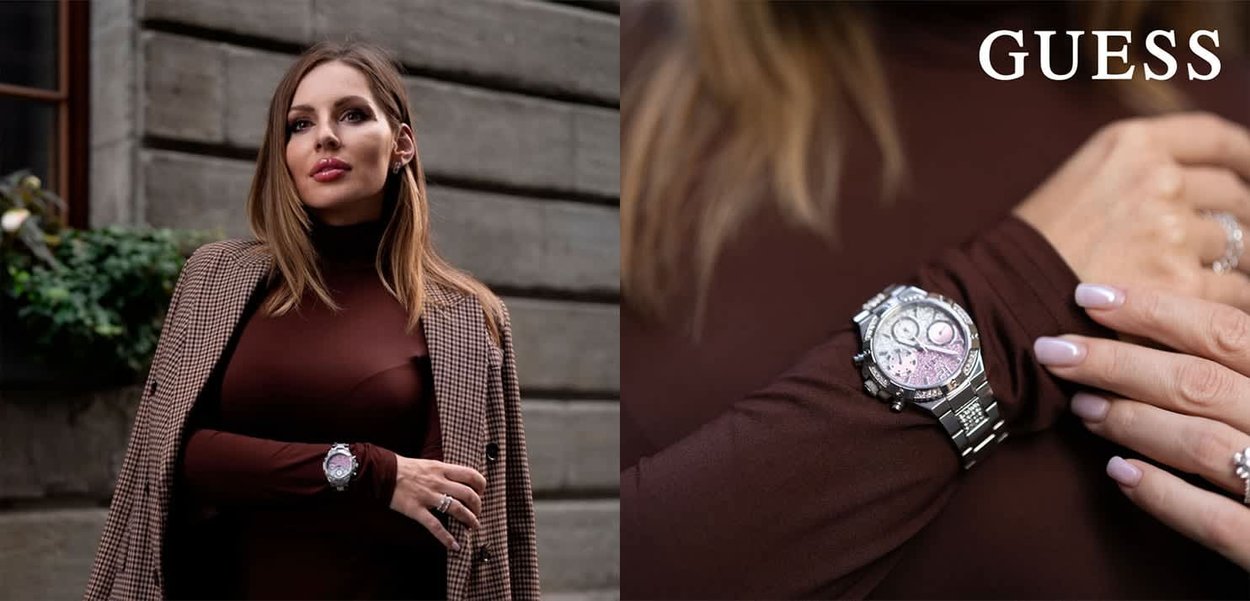 Reloj de acero inoxidable plateado con una esfera redonda, acentos rosas y una correa de eslabones metálicos, llevado sobre una blusa de manga larga marrón, con un abrigo a cuadros.