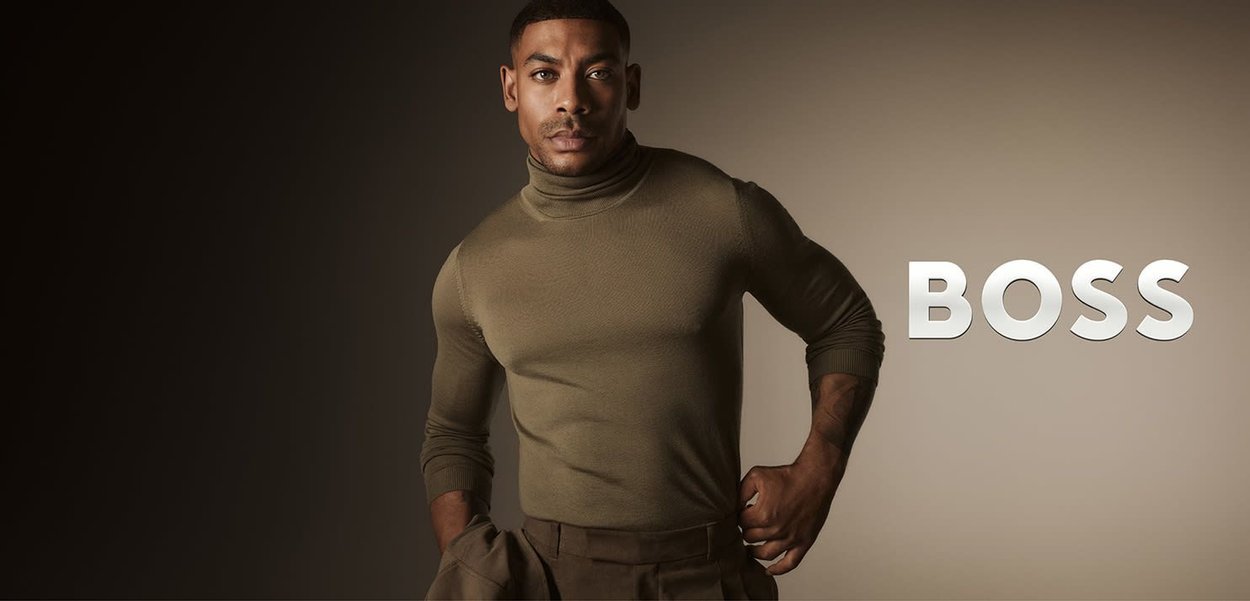 Olivgrüner Rollkragenpullover an einem männlichen Model, figurbetontes Design, getragen mit lockeren braunen Hosen, vor einem dunklen Farbverlauf-Hintergrund. "BOSS"-Schriftzug angezeigt.
