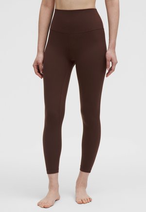 Nainen pukeutuneena tummanruskeisiin korkeavyötäröisiin kokonaan peittäviin legginsseihin seisomassa paljain jaloin yksiväristä taustaa vasten.