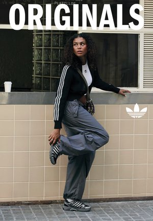 Cardigan nero corto con strisce bianche, pantaloni cargo grigi e sneakers con motivo bianco e nero. Sfondo con parete piastrellata.