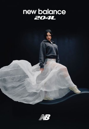 Vrouw in een wijdvallende witte rok en een donker sweatshirt poseert met witte New Balance 204L sneakers tegen een zwarte achtergrond.