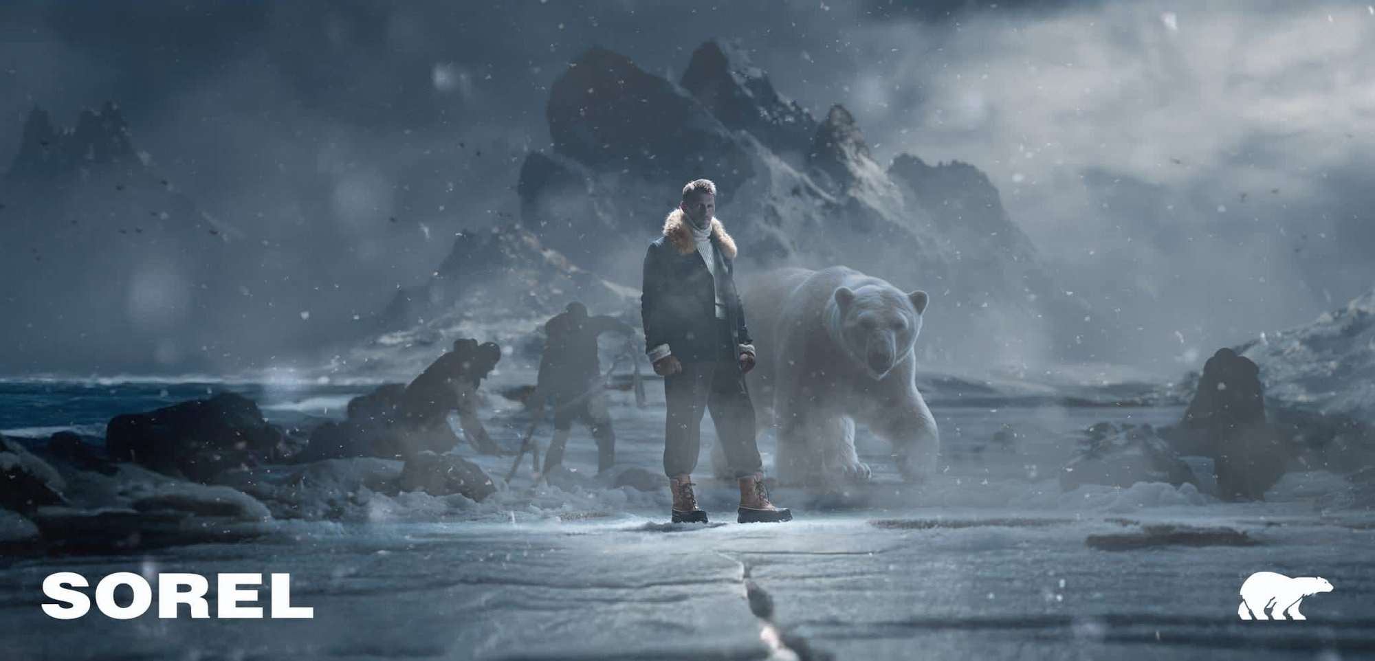 A person stands in snowy terrain wearing a dark jacket with fur trim and sturdy brown boots. A large polar bear is nearby, with mountains in the background.