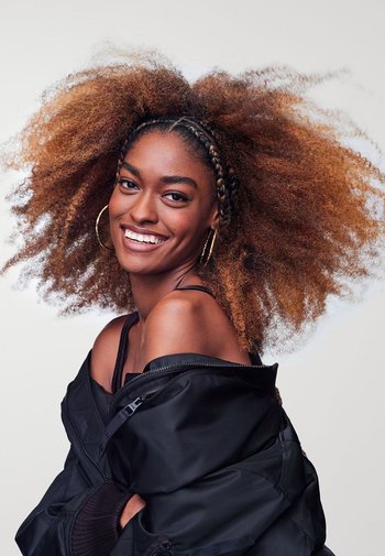 Black bomber jacket, off-shoulder, worn over a dark top. Hair is voluminous, curly, with braided sections. Gold hoop earrings accentuate the look.
