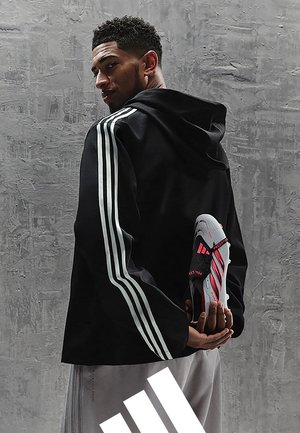 Mężczyzna w czarnej bluzie z kapturem z białymi paskami trzyma szaro-różowy but sportowy Adidas za plecami na tle szarej teksturowanej ściany.