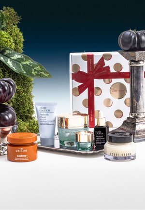 Set of skincare products on a silver tray: orange jar, various creams in translucent and opaque containers, decorative gift box, and dark fruit accents.