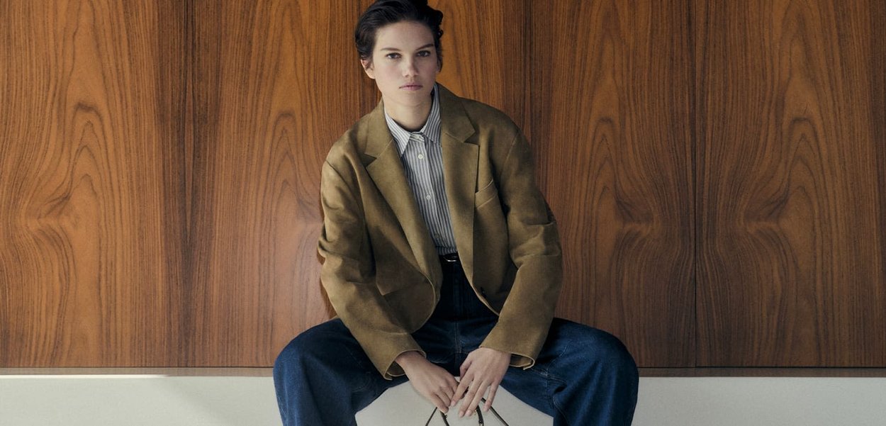 Brown suede blazer over a striped shirt, paired with dark blue wide-leg jeans. The backdrop features a textured wood panel.