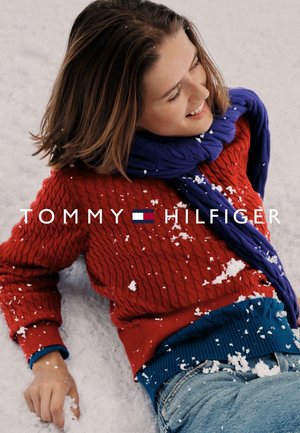 Pull rouge en maille torsadée, ceinture côtelée bleue et écharpe violette. Flocons de neige sur les vêtements. Mannequin allongé dans la neige. Logo bien visible.