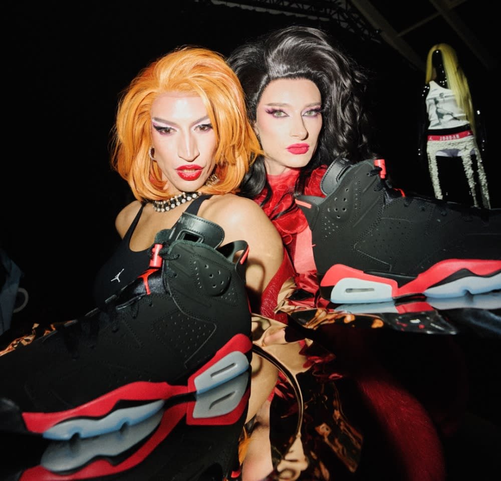 Dos personas con peinados y maquillaje posan detrás de zapatillas altas negras y rojas exhibidas sobre una superficie reflectante.