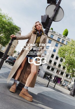 Femme blonde en long manteau beige et bottes UGG à plate-forme pose avec confiance dans une rue de la ville près d'un lampadaire.