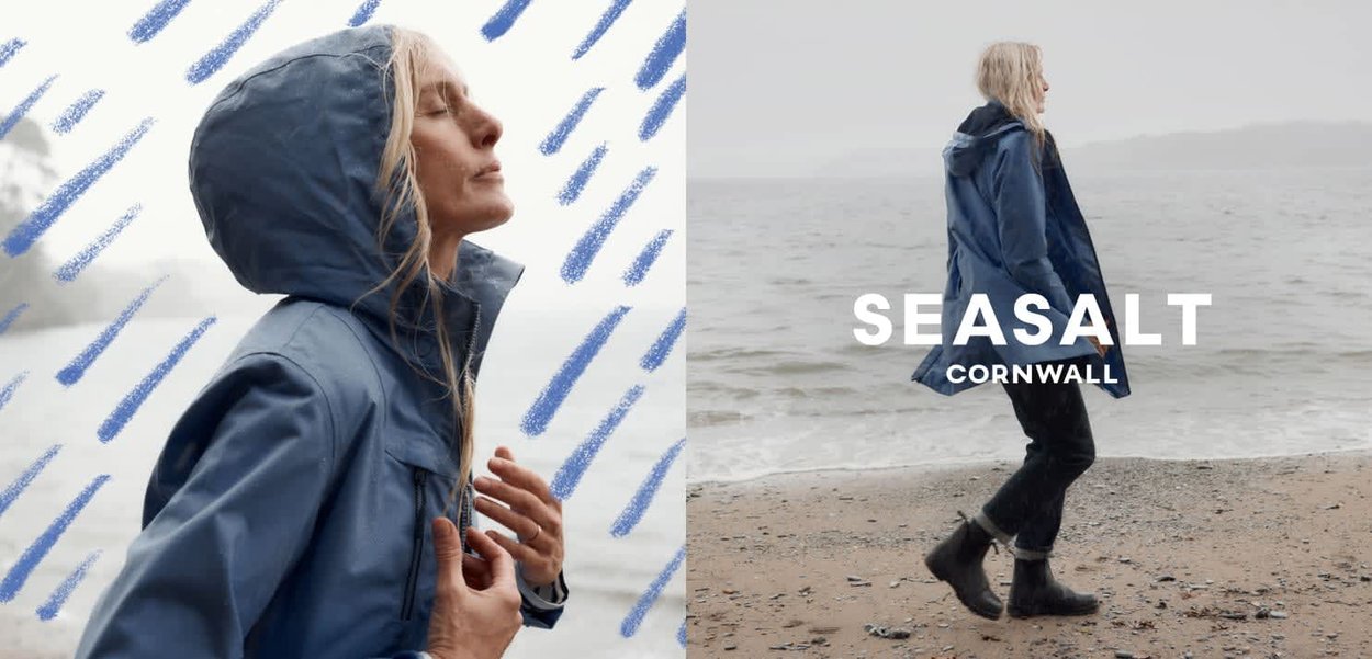 Femme portant une veste de pluie bleue et des bottes noires, debout sur une plage de galets, face à la mer sous un ciel couvert.