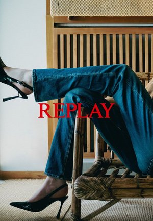 Mörkblå denimjeans med rak ben, kombinerat med svarta lacklädershögklackade skor. Foten vilar på en texturerad, vävd yta.