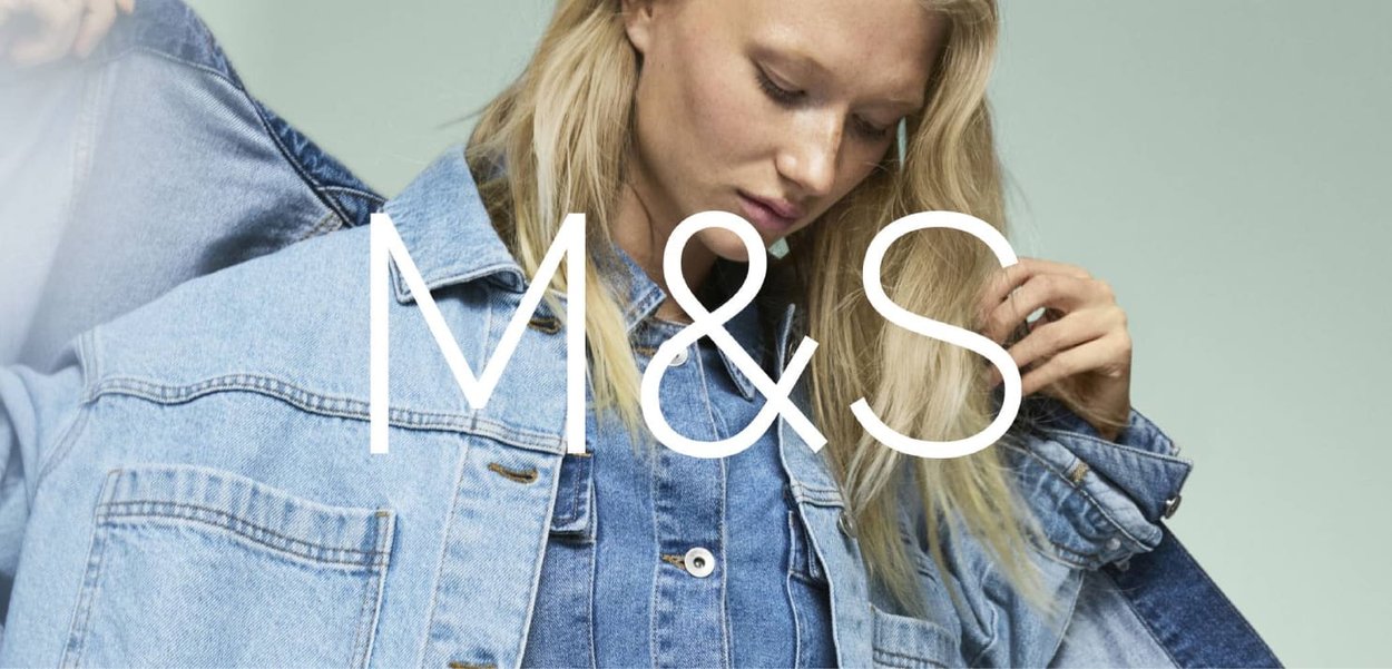 Jeune femme ajustant une veste en denim bleu clair sur une chemise en denim plus foncé, le tout sur un fond clair uni avec le logo M&S superposé.