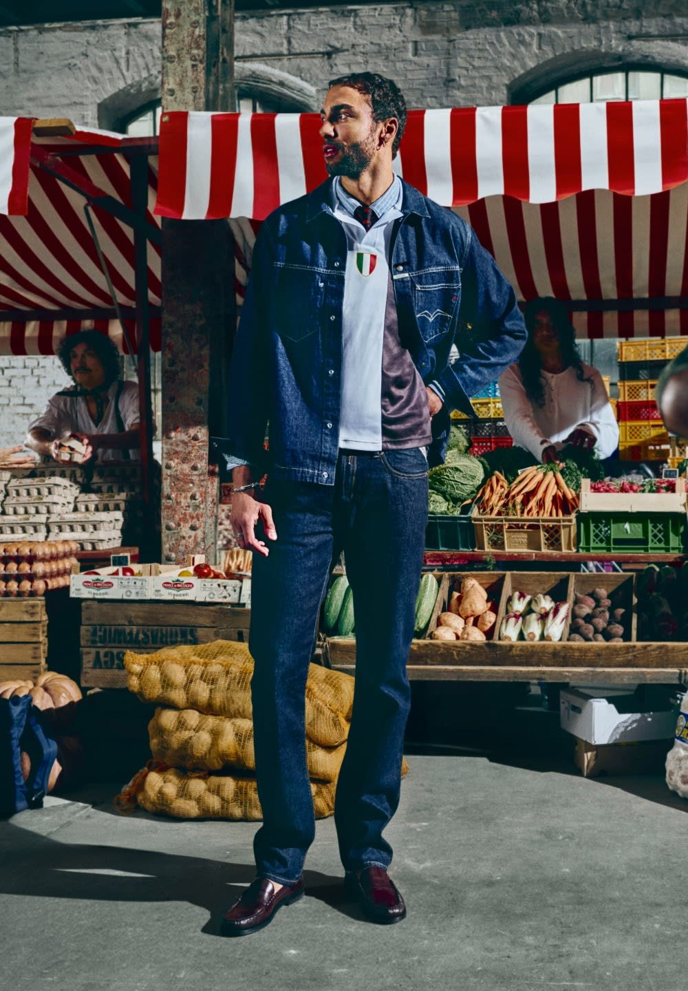 Giacca di jeans sopra una polo grigia e bianca con la bandiera italiana, jeans scuri e scarpe lucide. Ambientazione di un mercato con prodotti freschi sullo sfondo.