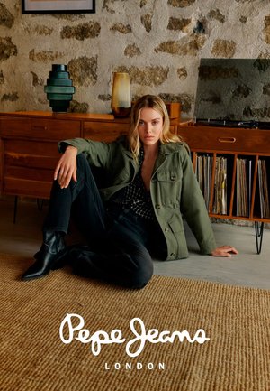 Junge Frau in grüner Jacke, schwarzen Hosen und Stiefeln sitzt auf dem Boden neben einem Holzsideboard mit Plattenspieler und Schallplatten.