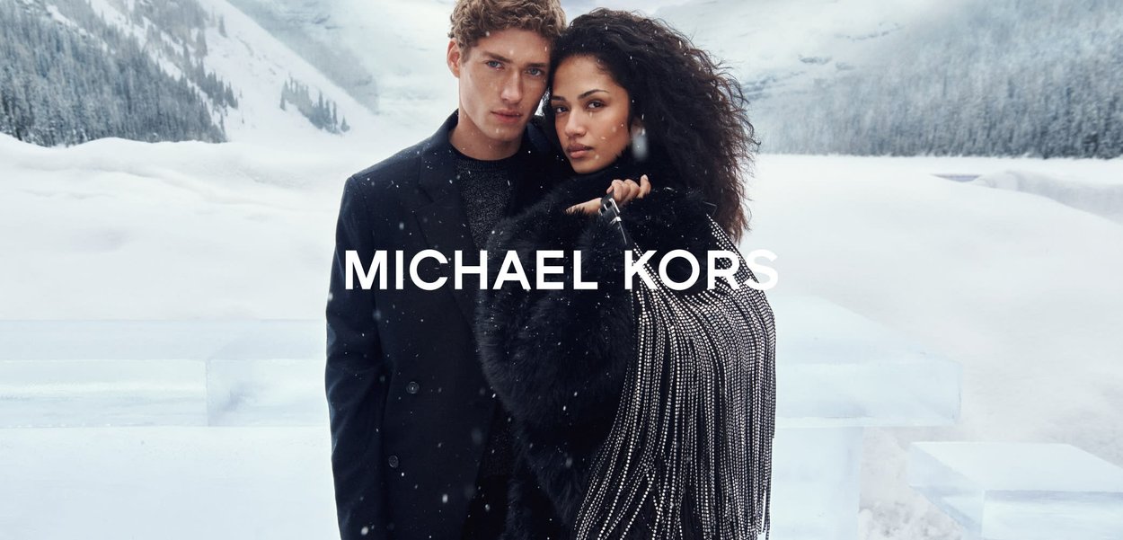 Elegante cappotto nero e sciarpa in pelliccia testurizzata con dettagli in cristallo, indossati da un uomo e una donna davanti a uno sfondo innevato, con la scritta "MICHAEL KORS".