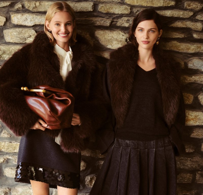 Twee modellen dragen donkere bontjassen; één houdt een diepe bruine leren clutch vast. De achtergrond toont natuurlijke steentexturen, wat het geheel versterkt.