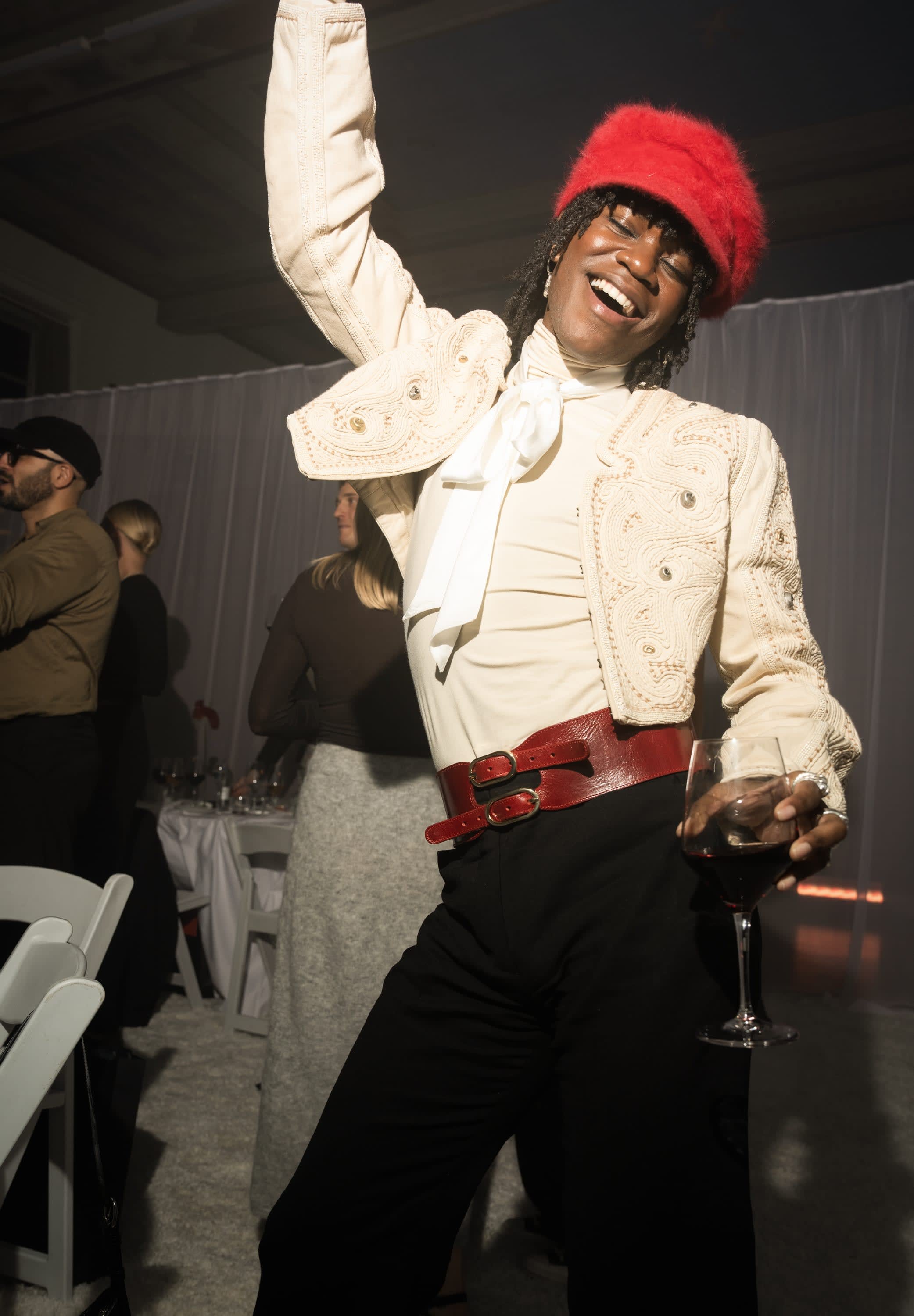 Person som bär en krämfärgad kort jacka med broderi, vit skjorta med rosett, rött läderbälte och håller i ett glas rödvin.