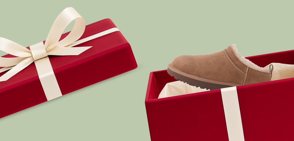 Scatola regalo rossa con un nastro crema, parzialmente aperta per rivelare una pantofola in pelle scamosciata marrone chiaro foderata in pelliccia sintetica, appoggiata su carta velina bianca.