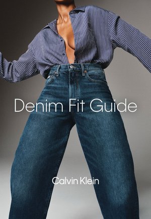 Person iført højtaljede vide denim jeans og en åben blåstribet skjorte, poserer med armene strakt ud mod en ensfarvet baggrund.
