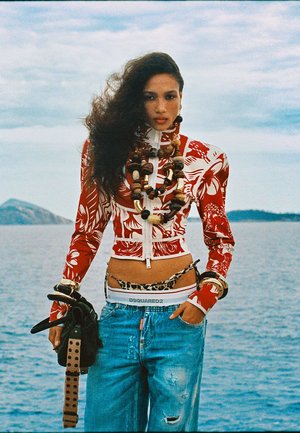 Mujer con cabello largo, oscuro y rizado lleva una chaqueta corta con patrón rojo y blanco, collares de cuentas de madera en capas, vaqueros desgastados y sostiene guantes junto al mar.