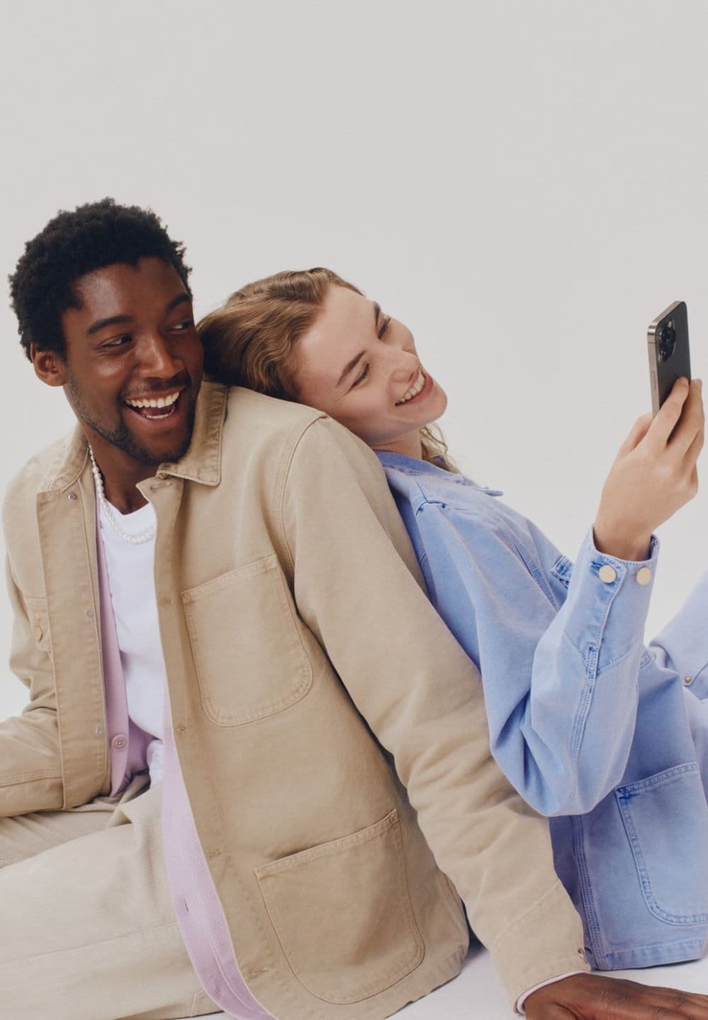 Du du žmonės sėdi vienas šalia kito, vilkėdami smėlio spalvos švarką ir šviesiai mėlyną džinsinį marškinį. Abu šypsosi, žiūrėdami į išmanųjį telefoną.