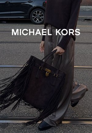 Grand sac à main en suède noir avec de longs détails en franges, accents en métal doré et mécanisme de verrouillage, porté à deux mains.
