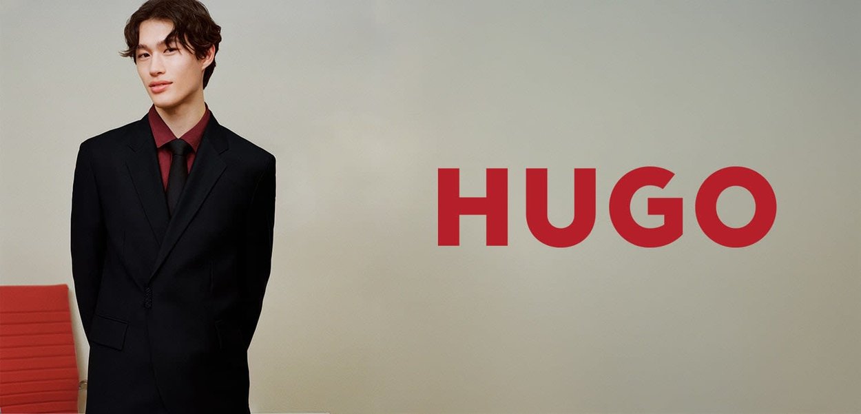 Costume noir avec revers crantés, associé à une chemise et une cravate bordeaux. En arrière-plan, une chaise rouge. Le nom de la marque "HUGO" est affiché en évidence.