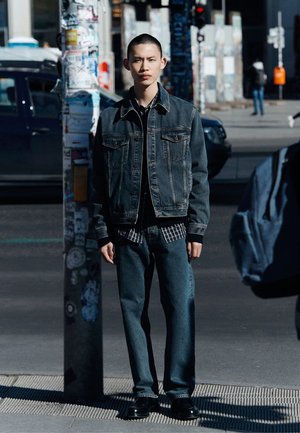 Mörk denimjacka lager över en svart skjorta, i kombination med matchande denimbyxor. Outfiten har en avslappnad passform och minimala detaljer.