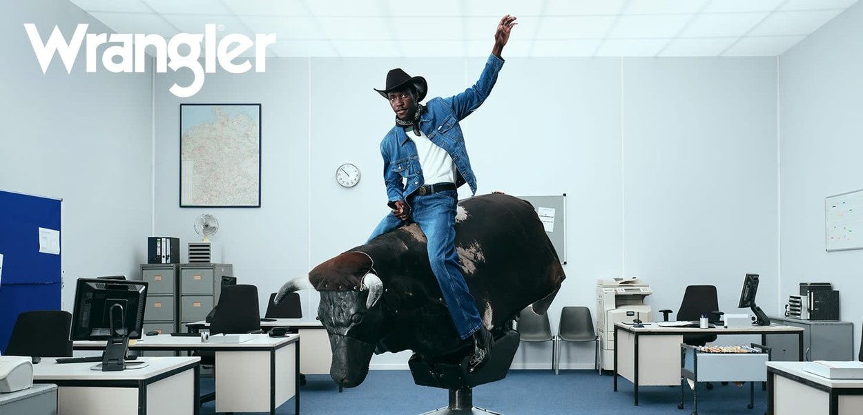 Cowboy auf einem mechanischen Bullen, trägt eine Jeansjacke, ein weißes Hemd und Jeans. Büroumgebung mit grauen Wänden, Computerarbeitsplätzen und Büromaterial.